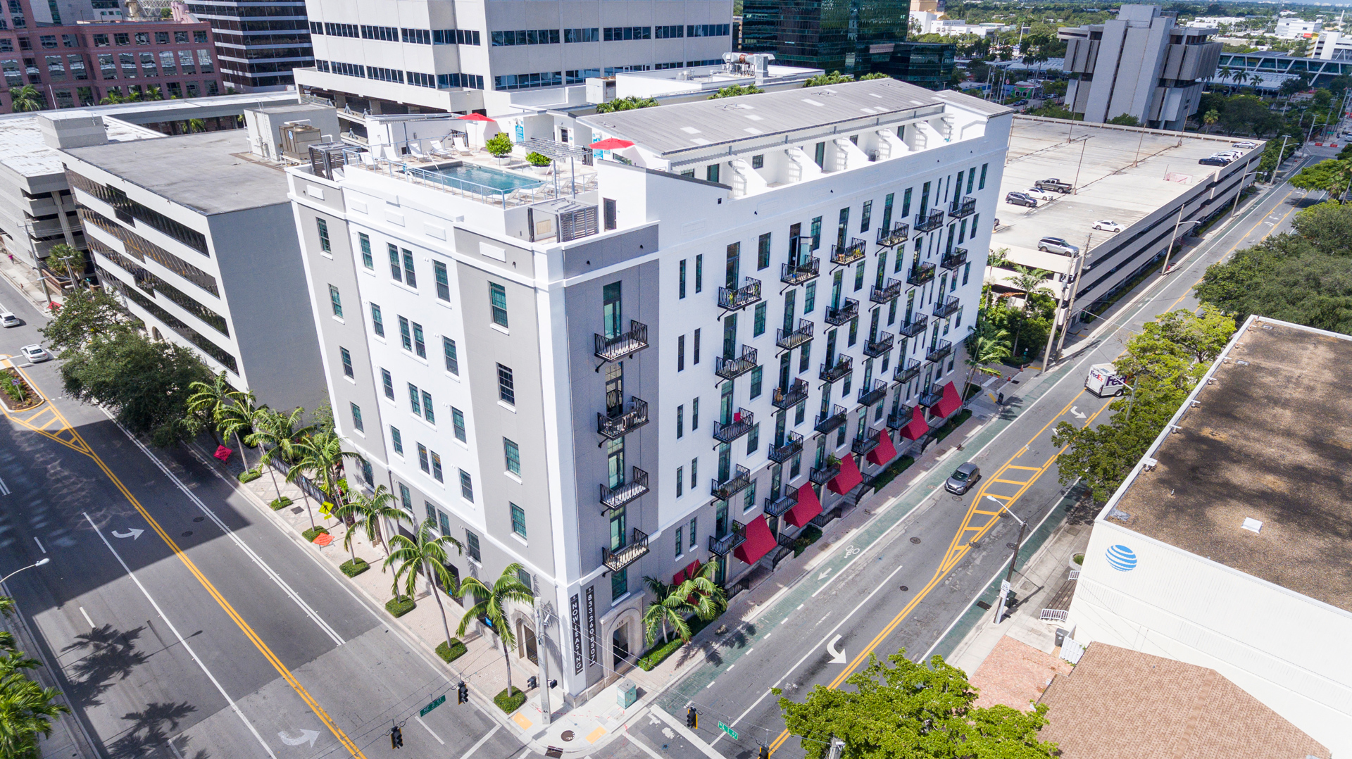 Aerial exterior view of The Exchange Lofts, a seven-story loft apartment building in downtown Fort Lauderdale with red balcony awnings, a rooftop pool, and palm-lined streets